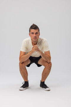 A young man squats low on a neutral grey background, looking at the camera with hands clasped. He wears a white tee and black shorts. Clean studio shot. Perfect for casual wear stock.