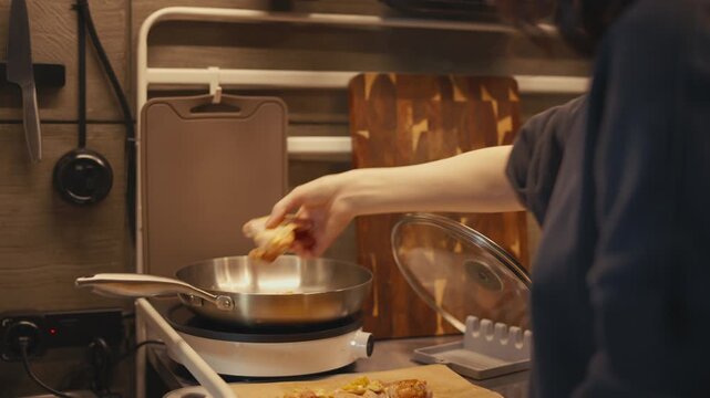 Side view of woman placing raw seasoned chicken wings into hot frying pan on induction stove in modern loft interior. Food prep and cooking process on stainless steel countertop in warm home kitchen.