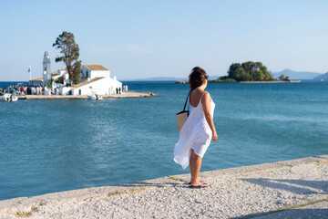 A woman in a white dress stands on a coastal pier in Kerkira, Greece, looking across the sea toward a whitewashed landmark. Clear blue waters surround the historic structure. © dsbedarieva
