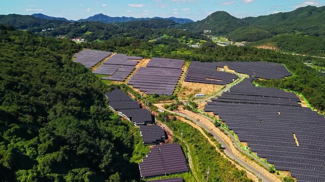 Aerial tracking left to right over terraced solar panel arrays carved into forested ridges in Gunbuk myeon Geumsan South Korea with service road and distant mountain village