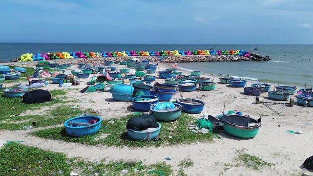 Low altitude drone perspective capturing a vibrant pattern of clustered traditional round fishing vessels and basket boats resting on the beach of Mui Ne, Vietnam with wave breaker background
