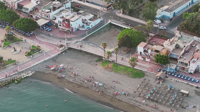 Aerial - urban beach in Malaga Spain with rows of sunbeds and parasols bathers on the sand a pedestrian footbridge over a stream mouth and a seafront promenade behind
