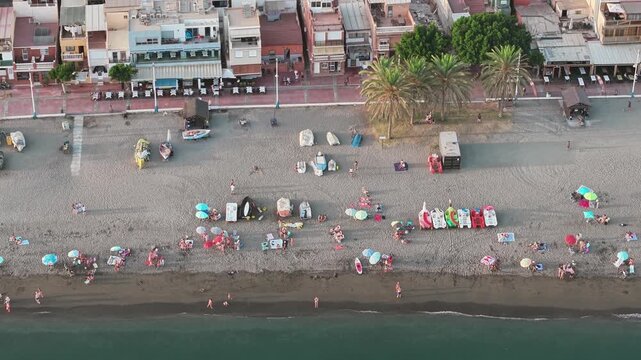 Top down aerial view of a busy urban beach in Malaga Spain with bathers parasols kayaks and small boats on the dark sand and a palm lined promenade of bars and apartments behind