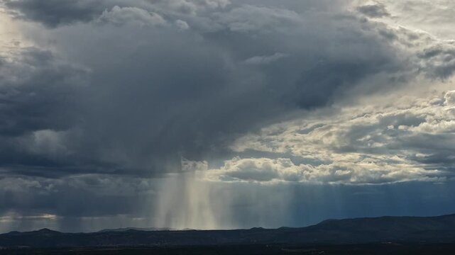 4K drone weather time lapse of a shimmering column of rain over a dark landscape. The sun perfectly breaks through a heavy cloud, intensely illuminating the falling precipitation.