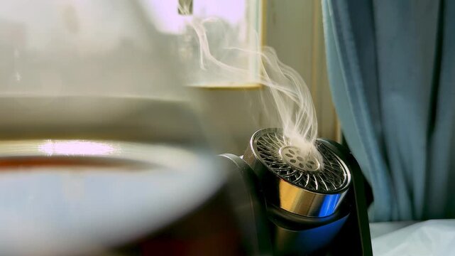 Close-up of a black electronic aroma diffuser emitting white smoke indoors. The device sits near a bright daylight window next to a blue curtain, with a blurred glass object in the foreground.