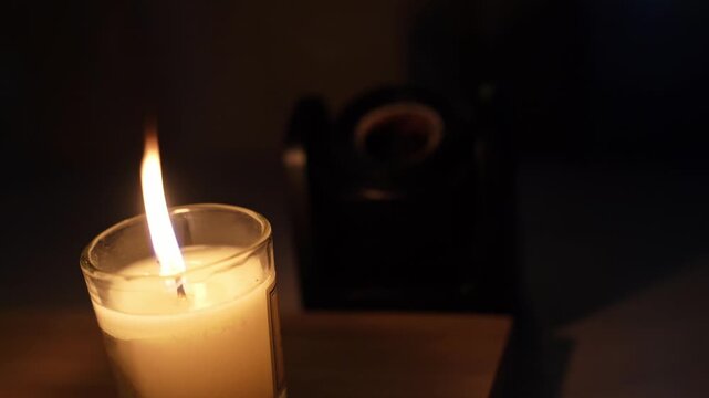 A close-up shot of a glowing candle burning inside a glass jar on a wooden surface in a dark room. The flame is suddenly blown out, plunging the scene into complete darkness.