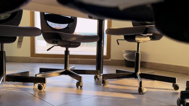 Low angle view from under a meeting desk showing empty rolling office chairs on a tiled floor. Warm daylight streams through a window, casting long shadows in the quiet corporate workspace.