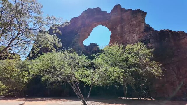 A rock arch rises above dense green trees with bright sun flare in this low angle view at Serra da Capivara National Park Brazil
