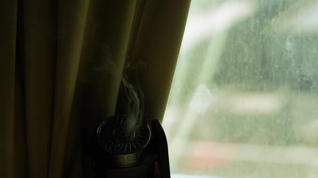 A close-up of a dark electronic aroma diffuser releasing a faint wisp of smoke or vapor. The small device sits indoors next to a bright, smudged window, partially hidden by a heavy shadowed curtain.