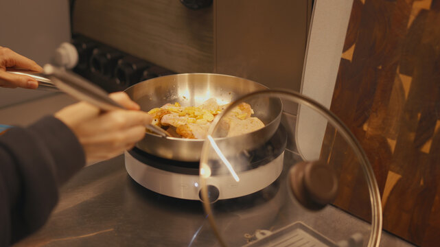 Close-up of woman hands turning chicken wings with a metal spatula in a stainless steel frying pan. Cooking poultry with ginger and garlic on a portable induction cooktop in a home kitchen.