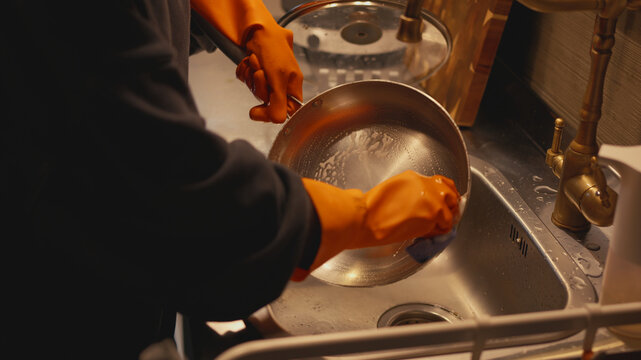 Close-up of a person in orange rubber gloves washing a metal frying pan using a soapy sponge in a stainless steel kitchen sink. A daily routine to keep the kitchen clean.