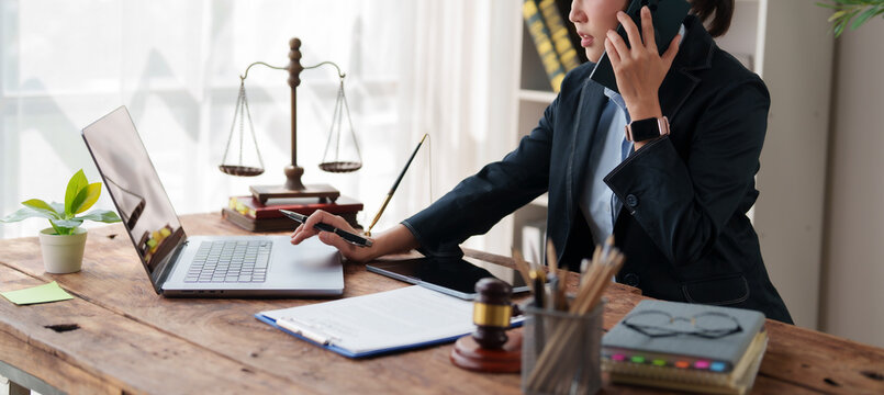 Female lawyer working remotely, discussing legal case on phone while using laptop in a professional home office, scales of justice and gavel on desk