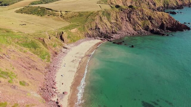 Drone orbit around a sandy cove, green pastures above red cliffs sweeping into clear sea, Pembrokeshire Coast National Park