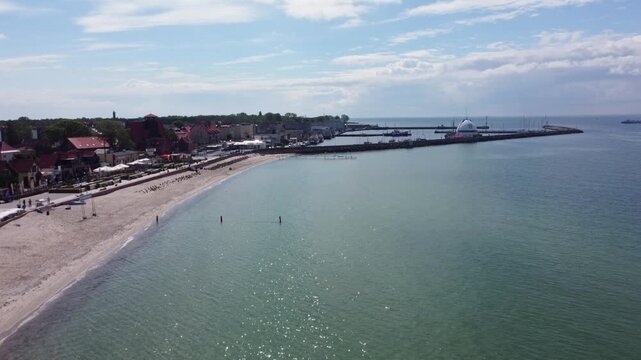 Flying toward the harbor, the drone moves over calm water along the sandy beach and coastal buildings of Hel in Poland.