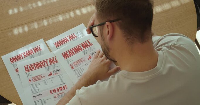 Shocked man looking at multiple expensive household bills, overwhelmed by rising living costs and financial pressure.