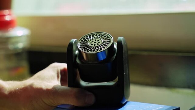 Close-up of a black portable electronic aroma diffuser emitting a thin trail of white vapor. The small device sits on a blue box indoors next to a bright window with a shallow depth of field.