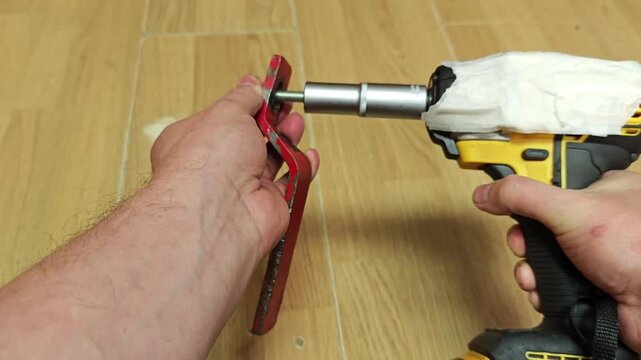 Impact driver and hex key handle red metal bracket on wood flooring. Hands steady piece while cordless tool backs out fastener, giving home repair work practical do it yourself feel.