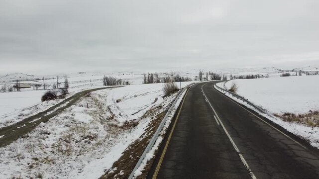 Aerial drone view: flight over a winding two-lane asphalt road with no overtaking traffic signs and an adjoining dirt road stretching through snow-covered rural fields on a cloudy winter day.
