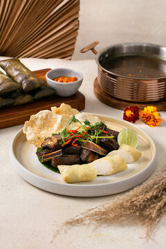 Indonesian braised beef ribs on banana leaf with rice cracker, steamed cassava, lime, and microgreens, served with lontong and sambal on wooden board.