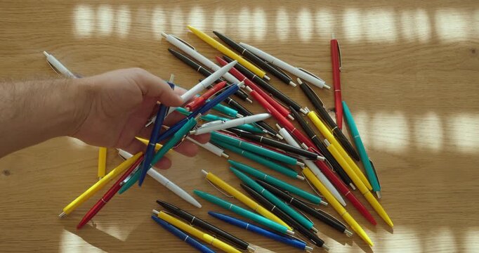 Close-up of hand sorting and picking colorful ballpoint pens from a large pile on a wooden table, representing choice, office supplies and stationery.
