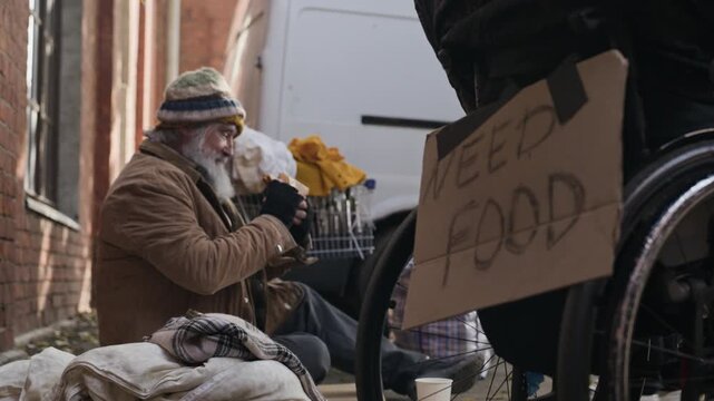 Senior Caucasian homeless man sitting near wheelchair with need food sign receives bread from volunteer and eats. Footage suitable for charity campaigns and social support advertising