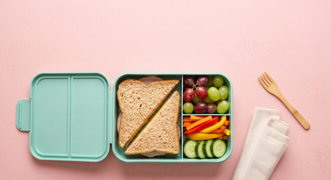 Lunchbox with sandwich, fruits, and vegetables on pink background