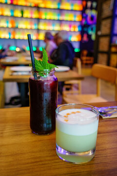 Two colorful drinks on a wooden restaurant table, including a berry beverage with mint and a creamy citrus drink, with diners blurred in the background