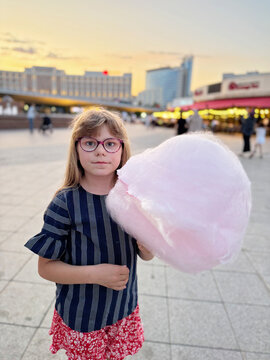 Young girl with glasses holding cotton candy at a city square during evening, childhood joy and summer fun captured in real life, real people