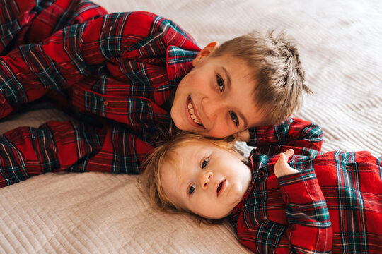 Two brothers lying together on a bed in matching red plaid shirts. Older boy and baby sibling sharing a warm family moment at home, representing brotherhood, childhood and family bonding