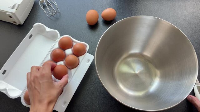 A hand takes a brown egg from a cardboard carton over a stainless steel bowl. The initial steps of baking or cooking preparation.
