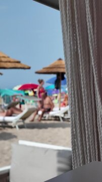 Vertical view from the shaded interior of the gazebo onto a sunny, sandy beach with thatched umbrellas, deck chairs and people relaxing in the blurry background. Empty space for text.