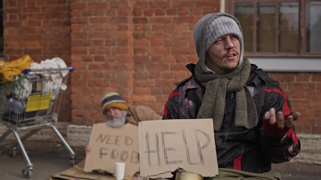 Young adult and senior Caucasian homeless men begging on street sidewalk holding help signs. Visual suitable for charity campaigns, social welfare documentaries, poverty awareness videos