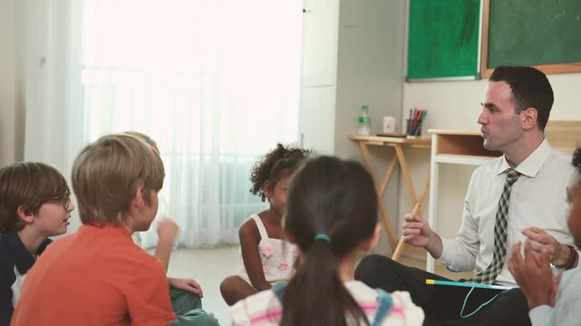 Male teacher leads elementary students sitting together in classroom circle activity, encouraging group discussion, participation, communication, collaborative learning.