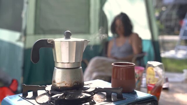 close up of moka pot italian coffee maker in the morning with smoke and warm light at camping site, woman in the background, lifestyle of camping life