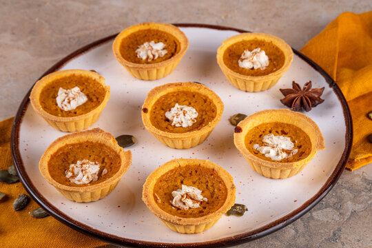 Sweet autumn baking, Mini pumpkin Tarts. Tiny pumpkin spice pies, with whipped cream topping and cinnamon powder, on beige concrete background