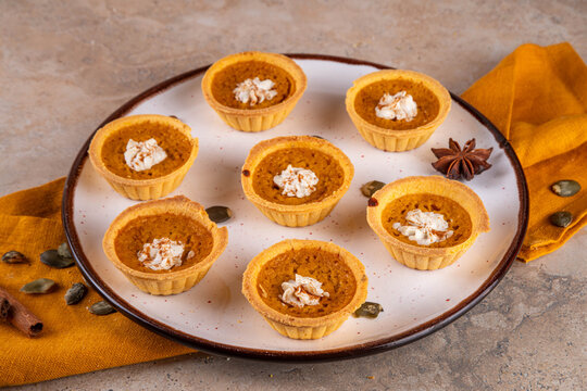 Sweet autumn baking, Mini pumpkin Tarts. Tiny pumpkin spice pies, with whipped cream topping and cinnamon powder, on beige concrete background