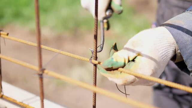Gloved hands twist thin wire around rusted rebar at construction site, holding steel bars steady during careful manual reinforcement work, with glove, hand, rebar, wire detail, showing construction