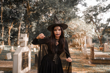 A mysterious witch in a black costume and pointed hat stands in a dark cemetery, surrounded by tombstones and deep shadows, creating an eerie Halloween atmosphere. © Kieng