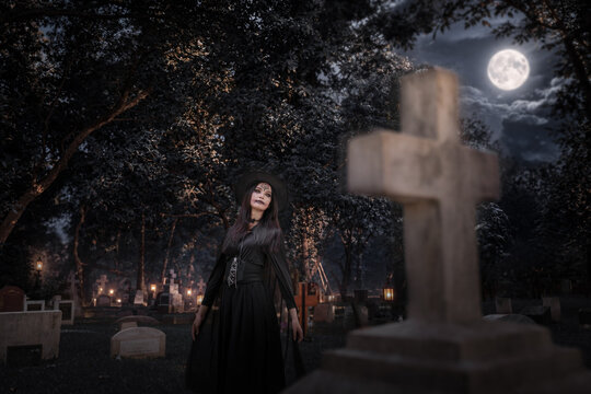 A mysterious witch in a dark cemetery under a full moon, surrounded by glowing lanterns and howling wolves, creating a spooky Halloween atmosphere.