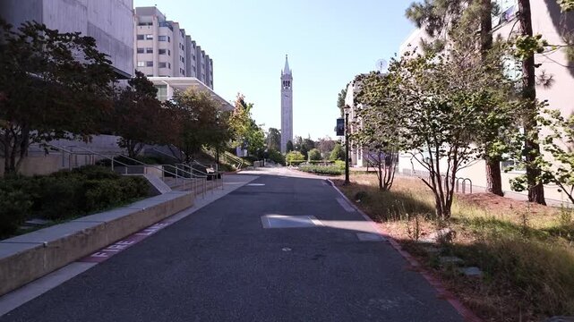 Personal perspective view walking on a paved pathway through the University of California Berkeley campus with the iconic Sather Tower, or Campanile, visible in the distance during a bright day