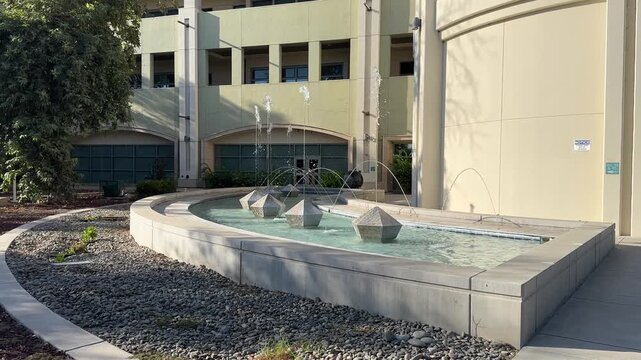 Wide shot of a contemporary water fountain at Modesto Junior College, featuring geometric stone sculptures, clear jets, and sunlight casting soft shadows on tranquil campus grounds.