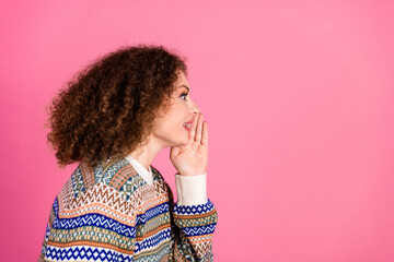 Young woman whispering a secret with a playful smile against a pink background wearing a colorful patterned sweater, side profile portrait conveying communication and friendly conversation © deagreez