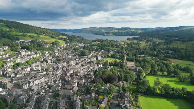 Aerial view of historic village beside tranquil lake. Grey stone homes cluster near green fields and hills. Ambleside town sits at the head of Windermere, in the lake district Cumbria UK