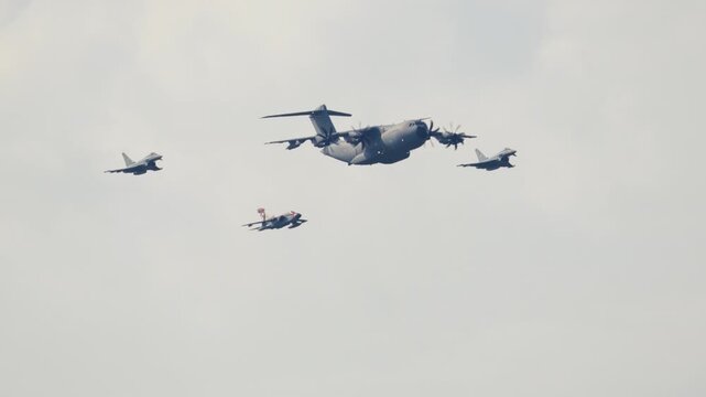 Formation of military aircraft flying together during an airshow. A large cargo plane is escorted by three smaller, faster fighter jets