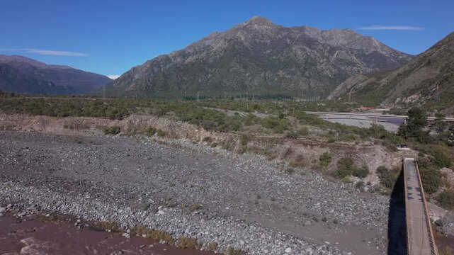 Ascending shot revealing wide rocky riverbed with sparse vegetation beneath towering rugged mountain peaks and a bridge crossing near a mountain highway