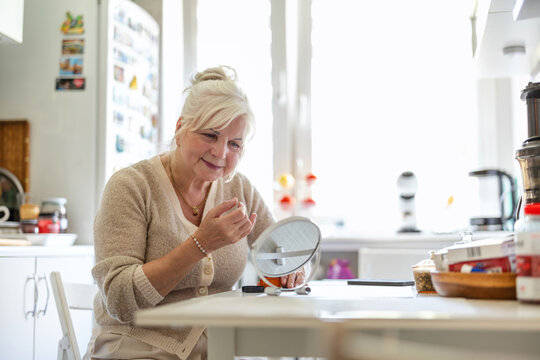 Senior woman using makeup mirror at the kitchen table
