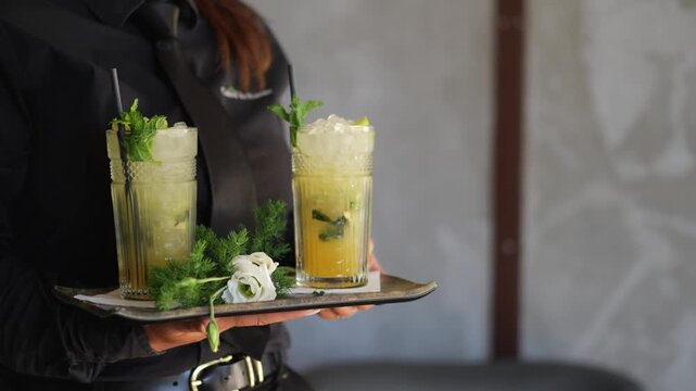 Waiter in black carries a tray with two tall cocktails garnished with mint and crushed ice toward guests at an outdoor wedding reception.
