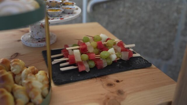 Watermelon melon and grape skewers arranged on a slate board beside savory pastries and custard tarts on a wooden catering table.