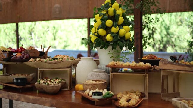 Tall lemon arrangement in a white vase rises above wooden bowls of appetizers and pastries on a buffet table beside a swimming pool.