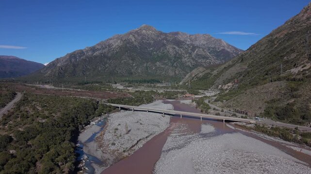 Orbit shot over a bridge crossing a reddish river flanked by rocky gravel bars beneath towering rugged peaks in a lush forested mountain valley